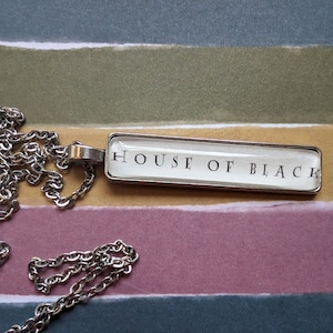 May include: A silver chain necklace with a rectangular pendant that says "HOUSE OF BLACK" in black lettering on a white background.
