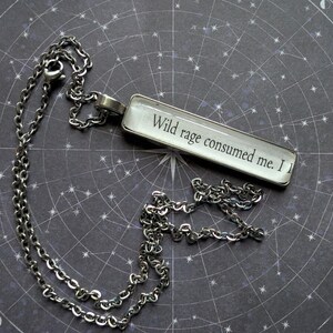 May include: Silver-toned necklace with a rectangular pendant. The pendant features the text "Wild rage consumed me. I" in a serif font against a white background. The chain and pendant are set against a dark background with a celestial design.