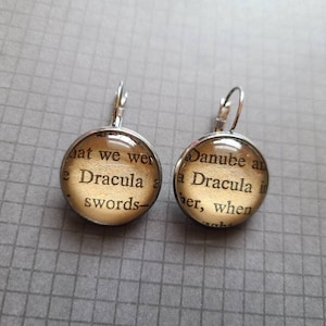 May include: A pair of silver dangle earrings with glass cabochons featuring text from the novel Dracula. The text on the left earring reads "hat we wer e Dracula a swords-" and the text on the right earring reads "Danube an a Dracula i er, when".