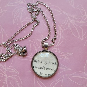 May include: A silver chain necklace with a round pendant featuring a white background with black text that reads "Brick by brick I wasn't enough r the way".