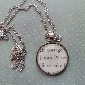 Puede incluir: Un collar de cadena de plata con un colgante redondo de vidrio que presenta un extracto impreso de un libro. El texto dice "their cottage. James Potter wife to take h"
