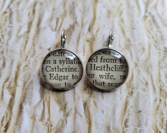 May include: A pair of silver dangle earrings with round glass charms. The charms feature text from the book "Wuthering Heights" by Emily Brontë. The text on the left charm reads "dare on a syllable Catherine. Edgar to pose to." The text on the right charm reads "ice ed from to Heathcliff," ur wife, m that reas"