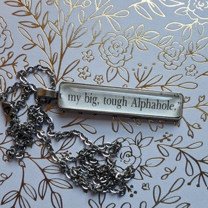 Puede incluir: Un collar plateado con un colgante rectangular. El colgante presenta el texto "... my big, tough Alphahole," en negro. La cadena del collar también es plateada y tiene un diseño de eslabones. El fondo es blanco con patrones florales dorados.