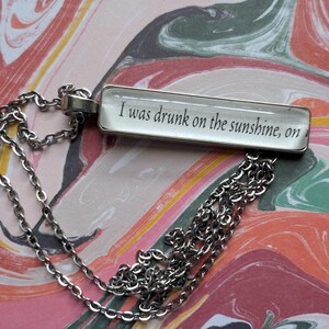 May include: Silver-toned necklace with a rectangular pendant. The pendant features the text "I was drunk on the sunshine, on" in black font. The background is a colorful, swirled pattern of pink, orange, and green.