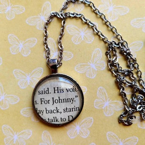 Puede incluir: Un collar plateado con un colgante redondo que muestra texto de un libro. El texto dice "said. His voic s. For Johnny." La cadena y el colgante están sobre un fondo amarillo con patrones de mariposas blancas.