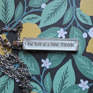 Puede incluir: Un collar de cadena de plata con un colgante rectangular de vidrio que dice "One foot at a time, friends."