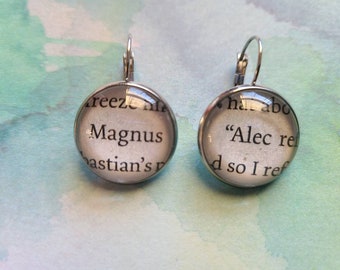 Puede incluir: Un par de pendientes de aro de plata con abalorios redondos de cristal. Los abalorios presentan texto de un libro. El abalorio izquierdo dice "Magnus" y el abalorio derecho dice "Alec re", "d so I ref".