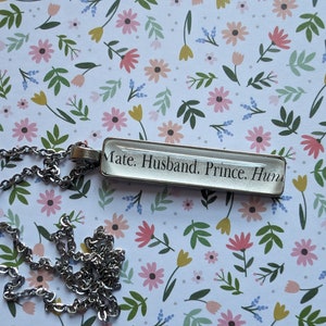 Puede incluir: Collar plateado con colgante rectangular. El colgante muestra el texto: "Mate. Husband. Prince. Hum". La cadena es de eslabones clásicos. El fondo es un estampado floral con flores rosas, amarillas y verdes.