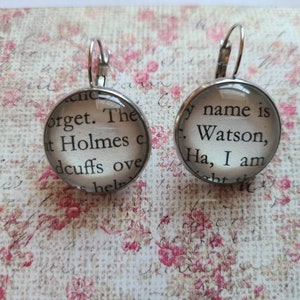 May include: A pair of round silver-tone leverback earrings. Each earring features a clear, domed glass cabochon with text from a book page. One earring reads "forget. The t Holmes c dcuffs ove hel" and the other reads "my name is Watson, Ha, I am".