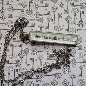 May include: A silver-toned necklace with a rectangular pendant. The pendant features the quote: "Also, I am wildly curious. I ha". The background is a pattern of antique keys.