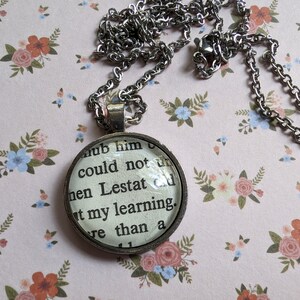 May include: A silver chain necklace with a round glass pendant featuring a black and white text excerpt from a book. The text reads "...hub him could not then Lestat out my learning...re than a".