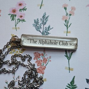 Puede incluir: Un collar de cadena de plata con un colgante rectangular de vidrio que dice "The Alphahole Club was".