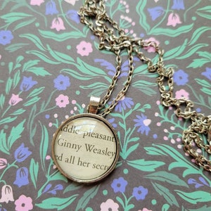 May include: A silver chain necklace with a round glass pendant featuring a printed excerpt from a book. The text reads "addle pleasant Ginny Weasley ed all her secr".