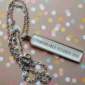 May include: A silver chain necklace with a rectangular pendant that says "Undesirable Number One." The pendant is white with black lettering. The necklace is on a brown background with colourful confetti.