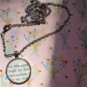 May include: A silver chain necklace with a round glass pendant featuring a printed excerpt from the book "Craigh na Dun" by Joe Abernathey. The text reads "Joe Abernathey Craigh na Dun approaching t out the hop".