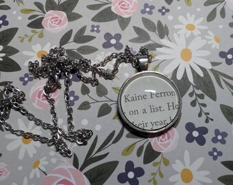 Könnte beinhalten: Eine silberfarbene Halskette mit Anhänger, die einen kreisförmigen Glasanhänger mit Text aus einem Buch zeigt. Der Text lautet "Kaine Ferron on a list". Die silberne Kette liegt auf einem floral gemusterten Hintergrund.