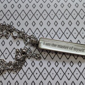 May include: A silver-toned chain necklace with a rectangular pendant. The pendant has the quote "I am the master of myself." in black text. The background is a white fabric with a black diamond pattern.