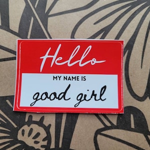 Könnte beinhalten: Ein rotes und weißes Namensschild mit dem Text "Hello" und "My name is good girl" in schwarzer Kursivschrift.