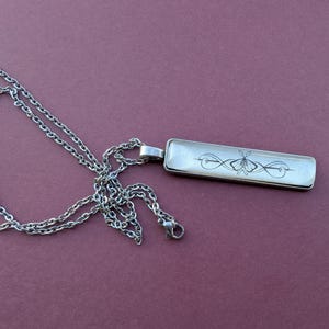 Op de afbeelding: Zilverkleurige ketting met een rechthoekige hanger. De hanger heeft een zwart-wit vlinderontwerp. De ketting is een klassieke schakelstijl. De achtergrond is een gedempt paars.