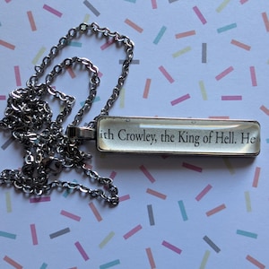 May include: Silver necklace with a rectangular pendant. The pendant displays the text "with Crowley, the King of Hell. He". The chain is a delicate link style, set against a background of pastel-colored shapes.