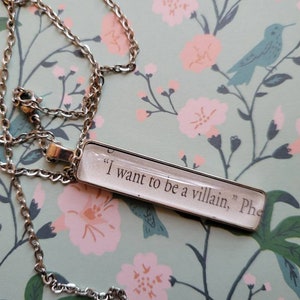 May include: A silver chain necklace with a rectangular pendant featuring the text "I want to be a villain, Phe"