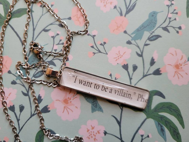 May include: A silver chain necklace with a rectangular pendant featuring the text "I want to be a villain, Phe"