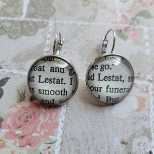 May include: A pair of silver hoop earrings with glass cabochons featuring text from the book "Interview with a Vampire" by Anne Rice. The text on the left earring reads "coat and g at Lestat. I s smooth and w", and the text on the right earring reads "e go. ad Lestat, s our funera But".