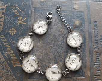 May include: A silver charm bracelet with six round glass charms. Each charm features a different quote from the book "Six of Crows" by Leigh Bardugo. The quotes are: "He's going to set Jesper straight against his d", "He met Lord. Nina held her hands", "With pi save the b", "Matthia is Wylan, better", "harpshe", "Inej wo ne weste", "rew up fr", "nal, Kaz wa", "tow".