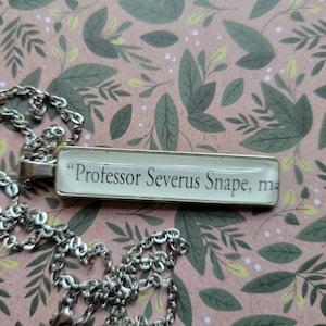 May include: A silver chain necklace with a rectangular pendant featuring the text "Professor Severus Snape, my"