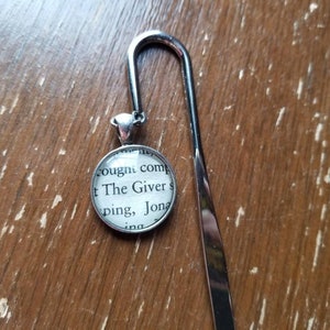 May include: A silver bookmark with a round charm featuring a book page with the text "The Giver" and a silver hook.