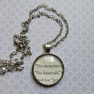 May include: A silver-toned necklace with a round pendant. The pendant features text from a book page, including the phrases "No mourners, No funerals." The chain is a classic link style, and the background is a light gray pattern.