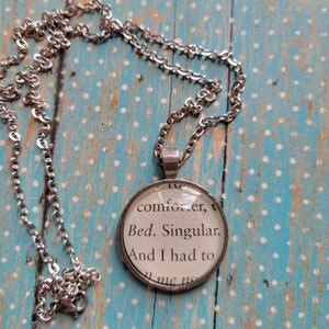 May include: A silver chain necklace with a round glass pendant featuring a printed quote: "To comforter, v. Bed. Singular. And I had to..."