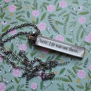 Puede incluir: Un collar de cadena de plata con un colgante rectangular que dice "there. Life was out there." El colgante está sobre un fondo con estampado floral.