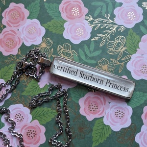 Puede incluir: Un collar de cadena de plata con un colgante rectangular que dice "certified Starborn Princess."