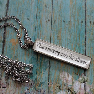 Puede incluir: Collar plateado con colgante rectangular. El colgante muestra el texto "What a fucking mess this all was" sobre un fondo blanco. El collar descansa sobre una superficie turquesa con estampado floral.