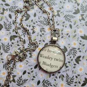 Puede incluir: Un collar de cadena plateada con un colgante redondo de vidrio que presenta una cita de la serie de Harry Potter: "their brooms. Weasley twins & Bludgers"