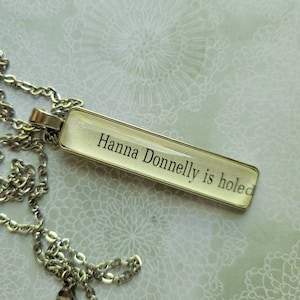 Puede incluir: Un collar de cadena plateada con un colgante de vidrio transparente que dice "Hanna Donnelly is holed".