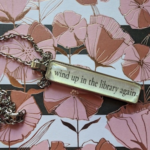 May include: A silver-toned necklace with a rectangular pendant. The pendant has the text "I wind up in the library again." The necklace chain is a delicate link style. The background features a floral pattern in pink, white, and black.