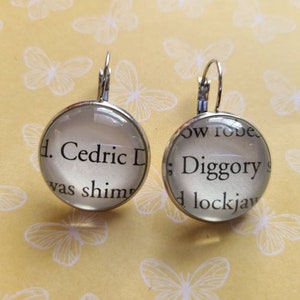 May include: A pair of silver earrings with glass charms. The charms feature text from the book "Harry Potter and the Goblet of Fire" by J.K. Rowling. The text on the left charm reads "Cedric D. was shimm", and the text on the right charm reads "ow robes: Diggory s d lockjaw".