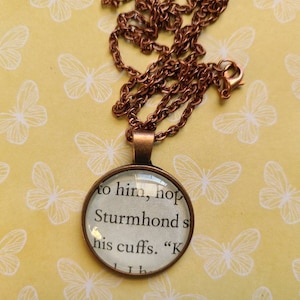 Puede incluir: Un collar de cadena de cobre con un colgante de vidrio que presenta una cita del libro "The Stormlight Archive" de Brandon Sanderson. La cita dice: "to him, hope Sturmhond's his cuffs."
