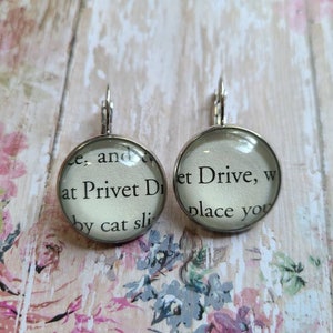 Puede incluir: Un par de pendientes de aro de plata con colgantes de cristal. Los colgantes presentan texto del libro "Harry Potter y la piedra filosofal" que dice "...y dos en Privet Drive, w...et Drive, w...place you...by cat sli..."