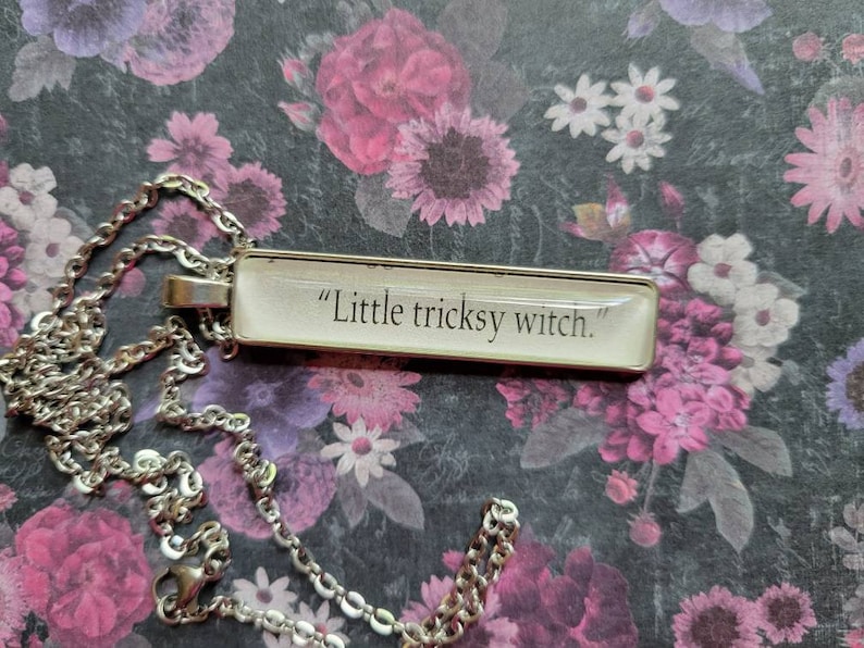 Op de afbeelding: Een zilveren ketting met een rechthoekige hanger met de tekst "Little tricksy witch."