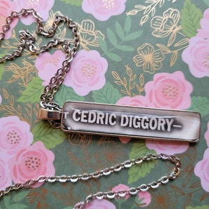 Peut inclure: Un collier en chaîne argent avec un pendentif rectangulaire qui dit "Cedric Diggory" en lettres noires.