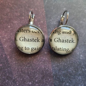May include: A pair of silver earrings with glass charms. The charms feature a white background with black text that reads "Asters of 1, Ghastek a ng to gair" and "Ang and p Ghastek. ulating, a".