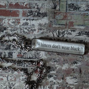 May include: A silver-toned necklace with a rectangular pendant. The pendant has the text "Whiners don't wear black." The background is a brick wall with a black and white toile pattern.