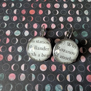 May include: A pair of silver-toned leverback earrings. Each earring features a round, clear glass cabochon with text from a book page. The background is a dark gray pattern with pink and teal crescent moon shapes.