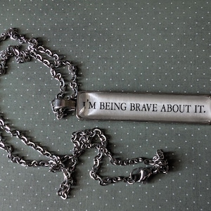 May include: A silver-toned necklace with a rectangular pendant. The pendant has the phrase "I'M BEING BRAVE ABOUT IT." printed in black on a white background. The necklace chain is a linked design.