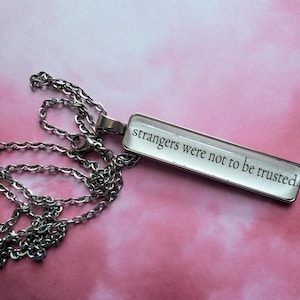 May include: Silver-toned necklace with a rectangular pendant. The pendant has the text "strangers were not to be trusted" in black font. The chain is a classic link style, and the background is a pink gradient.