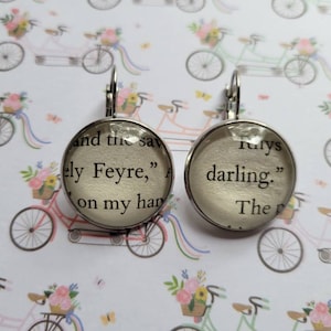 May include: A pair of silver earrings with glass cabochons featuring text from the book A Court of Thorns and Roses. The text on the left earring reads "and the sav ely Feyre," and the text on the right earring reads "rulys darling."