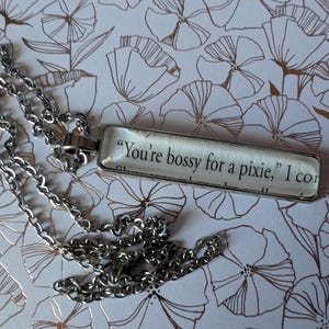 May include: A silver-toned necklace with a rectangular pendant. The pendant has the quote "You're bossy for a pixie, I con" printed on it. The necklace is displayed on a floral patterned background.